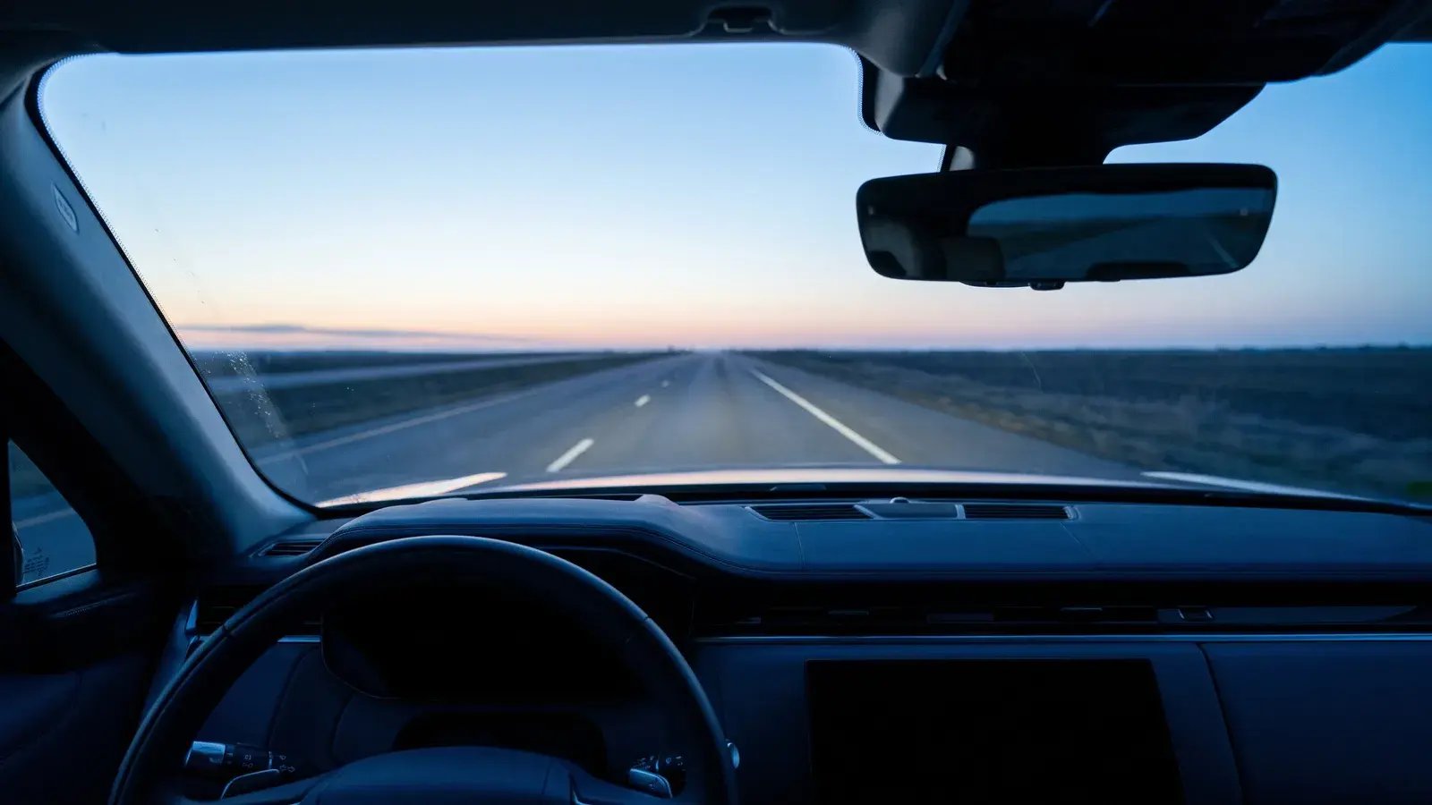 View from the driver’s seat through a windshield onto an open highway at dawn, the rearview mirror showing the road already travelled
